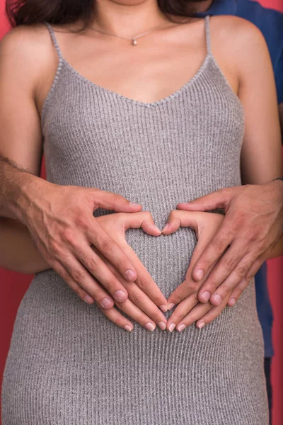 赤の背景に分離された妊娠中の妻の腹に自分の手で心を示す幸せな若いカップル — ストック写真