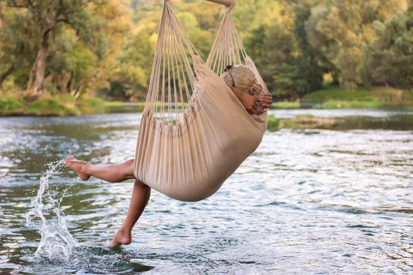 川のほとりで自然を楽しみながらハンモックで休んで若いブロンドの女性 — ストック写真
