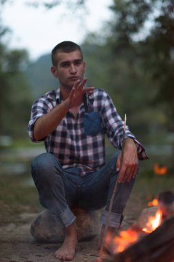 Bir grup mutlu genç arkadaş nehir kıyısında kamp ateşi etrafında dinleniyor ve eğleniyorlar.