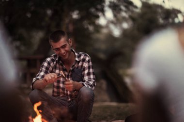 Bir grup mutlu genç arkadaş nehir kıyısında kamp ateşi etrafında dinleniyor ve eğleniyorlar.