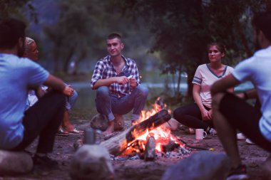 Bir grup mutlu genç arkadaş nehir kıyısında kamp ateşi etrafında dinleniyor ve eğleniyorlar.
