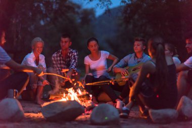 Bir grup mutlu genç arkadaş nehir kıyısında kamp ateşi etrafında dinleniyor ve eğleniyorlar.