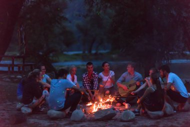 Bir grup mutlu genç arkadaş nehir kıyısında kamp ateşi etrafında dinleniyor ve eğleniyorlar.