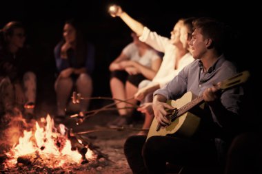 Bir grup mutlu genç arkadaş nehir kıyısında kamp ateşi etrafında dinleniyor ve eğleniyorlar.