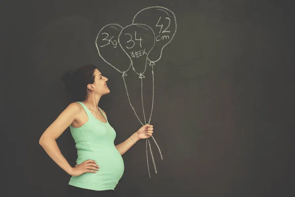 黒い黒板の前で腹に手を持つ幸せな妊娠中の女性の肖像画 — ストック写真