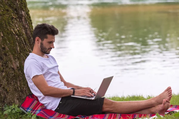 一个年轻的自由职业者 在河岸上的树下 在美丽的大自然下工作时 使用笔记本电脑 — 图库照片