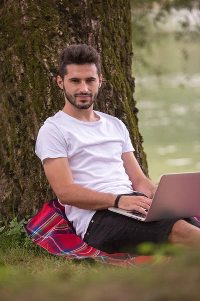 一个年轻的自由职业者 在河岸上的树下 在美丽的大自然下工作时 使用笔记本电脑 — 图库照片
