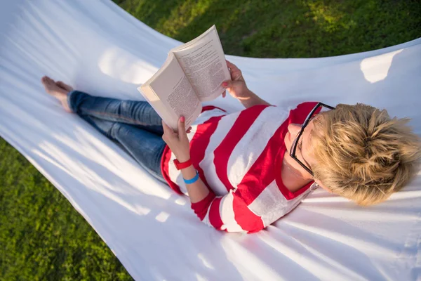 休暇中に静かな庭園でハンモックでリラックスしながら本を読む若い女性 — ストック写真