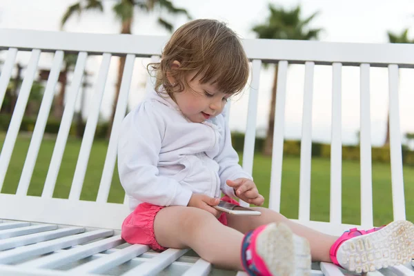 美丽的小女孩玩手机 而坐在公园的长凳上 — 图库照片