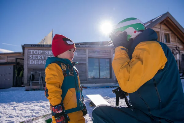 在阳光明媚的冬日里 年轻快乐的父亲在美丽的滑雪场里第一次在滑雪板上为小儿子做准备 — 图库照片