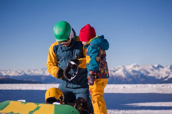 在阳光明媚的冬日里 年轻快乐的父亲在美丽的滑雪场里第一次在滑雪板上为小儿子做准备 — 图库照片