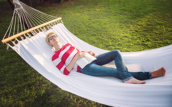 休暇中に静かな庭園でハンモックでリラックスしながら本を読む若い女性 — ストック写真