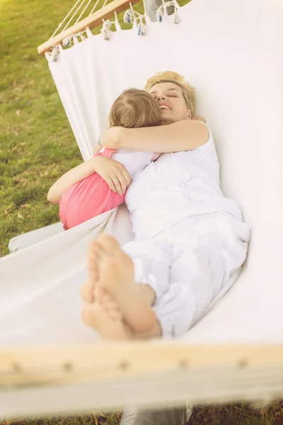 在度假屋花园的一个阳光明媚的日子里 快乐的母亲和一个小女儿享受着在吊床里拥抱和放松的空闲时间 — 图库照片