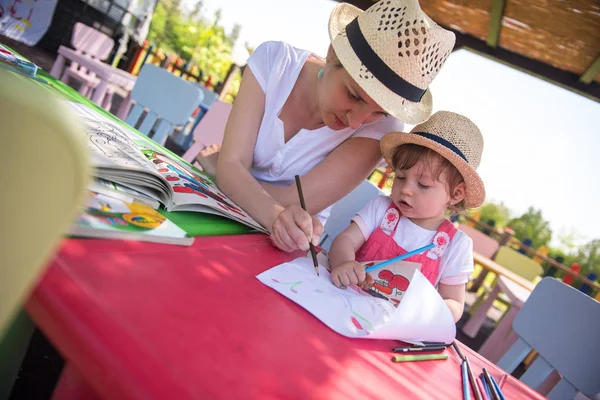 年轻的母亲和她的小女儿愉快地在一起 一边用铅笔蜡笔 一边在外面的游戏学校画一幅五颜六色的画 — 图库照片
