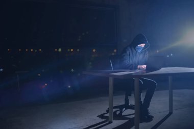 Genç yetenekli hacker karanlık ofiste çalışırken dizüstü bilgisayar kullanıyor. Arka planda büyük şehir ışıkları var.
