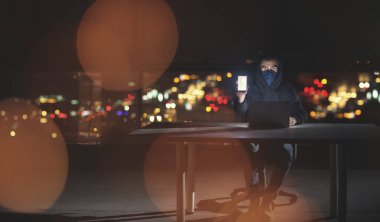 Genç yetenekli hacker karanlık ofiste çalışırken dizüstü bilgisayar kullanıyor. Arka planda büyük şehir ışıkları var.