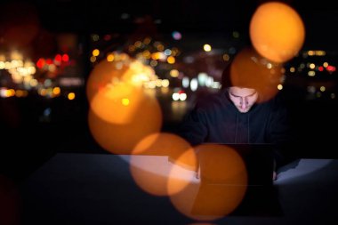 Genç yetenekli hacker karanlık ofiste çalışırken dizüstü bilgisayar kullanıyor. Arka planda büyük şehir ışıkları var.