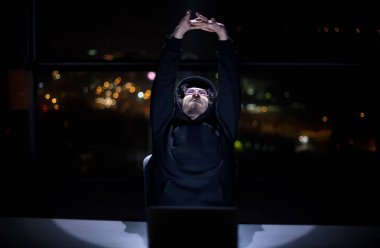 Genç yetenekli hacker karanlık ofiste çalışırken dizüstü bilgisayar kullanıyor. Arka planda büyük şehir ışıkları var.
