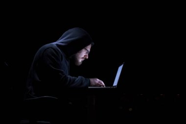 Genç yetenekli hacker karanlık ofiste çalışırken dizüstü bilgisayar kullanıyor. Arka planda büyük şehir ışıkları var.