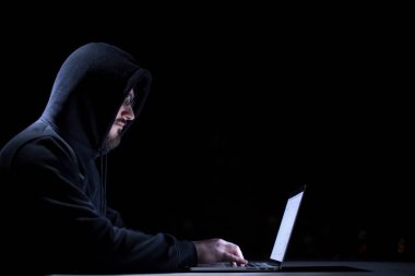 Genç yetenekli hacker karanlık ofiste çalışırken dizüstü bilgisayar kullanıyor. Arka planda büyük şehir ışıkları var.