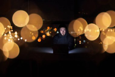 Genç yetenekli hacker karanlık ofiste çalışırken dizüstü bilgisayar kullanıyor. Arka planda büyük şehir ışıkları var.