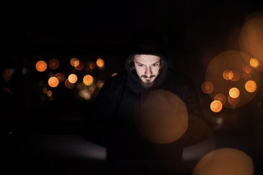 Genç yetenekli hacker karanlık ofiste çalışırken dizüstü bilgisayar kullanıyor. Arka planda büyük şehir ışıkları var.