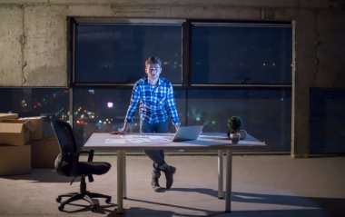 İnşaat alanındaki genç erkek mühendisin portresi gece yeni açılan ofiste dizüstü bilgisayarla belge ve iş akışını kontrol ediyor.