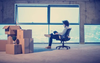 İnşaat alanındaki genç erkek mimar yeni açılan ofiste yanında karton kutularla dizüstü bilgisayar kullanarak belge ve iş akışını kontrol ediyor.