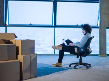 İnşaat alanındaki genç erkek mimar yeni açılan ofiste yanında karton kutularla dizüstü bilgisayar kullanarak belge ve iş akışını kontrol ediyor.