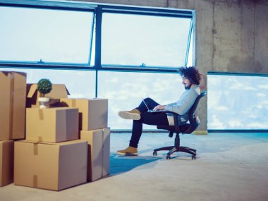 İnşaat alanındaki genç erkek mimar yeni açılan ofiste yanında karton kutularla dizüstü bilgisayar kullanarak belge ve iş akışını kontrol ediyor.