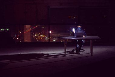 Genç yetenekli hacker karanlık ofiste çalışırken dizüstü bilgisayar kullanıyor. Arka planda büyük şehir ışıkları var.
