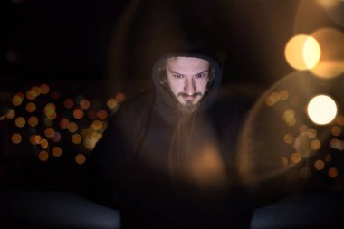 Genç yetenekli hacker karanlık ofiste çalışırken dizüstü bilgisayar kullanıyor. Arka planda büyük şehir ışıkları var.