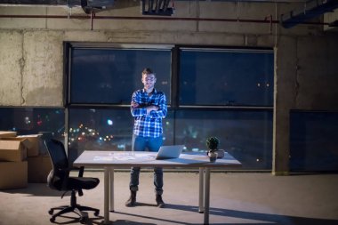 İnşaat alanındaki genç erkek mühendisin portresi gece yeni açılan ofiste dizüstü bilgisayarla belge ve iş akışını kontrol ediyor.