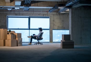 İnşaat alanındaki genç erkek mimar yeni açılan ofiste yanında karton kutularla dizüstü bilgisayar kullanarak belge ve iş akışını kontrol ediyor.