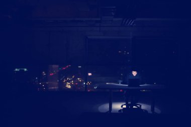 Genç yetenekli hacker karanlık ofiste çalışırken dizüstü bilgisayar kullanıyor. Arka planda büyük şehir ışıkları var.
