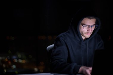 Genç yetenekli hacker karanlık ofiste çalışırken dizüstü bilgisayar kullanıyor. Arka planda büyük şehir ışıkları var.