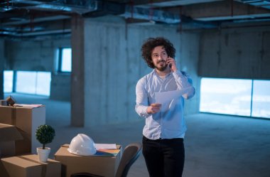 İnşaat alanındaki genç erkek mimar, yeni açılan ofiste etrafında karton kutular olan bir cep telefonu kullanarak belge ve iş akışını kontrol ediyor.