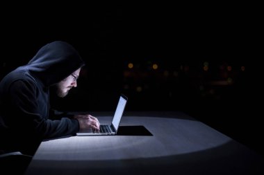 Genç yetenekli hacker karanlık ofiste çalışırken dizüstü bilgisayar kullanıyor. Arka planda büyük şehir ışıkları var.