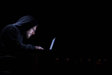 Genç yetenekli hacker karanlık ofiste çalışırken dizüstü bilgisayar kullanıyor. Arka planda büyük şehir ışıkları var.