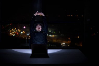 Genç yetenekli hacker karanlık ofiste çalışırken dizüstü bilgisayar kullanıyor. Arka planda büyük şehir ışıkları var.