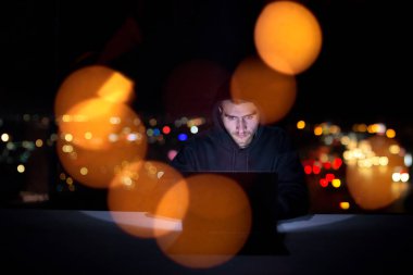 Genç yetenekli hacker karanlık ofiste çalışırken dizüstü bilgisayar kullanıyor. Arka planda büyük şehir ışıkları var.