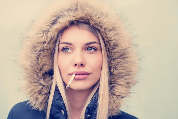 若いですブロンドの女の子身に着けています冬のジャケットフードとタバコで口の中に隔離された白い背景 — ストック写真