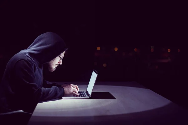 夜に大都市の照明を背景に暗いオフィスで作業しながら ラップトップコンピュータを使用して若い才能のハッカー — ストック写真