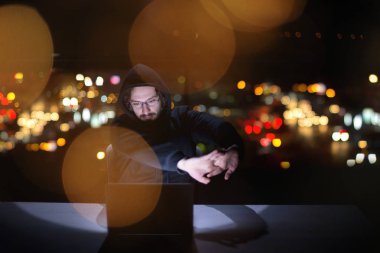 Genç yetenekli hacker karanlık ofiste çalışırken dizüstü bilgisayar kullanıyor. Arka planda büyük şehir ışıkları var.