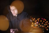 Mladý talentovaný hacker pomocí notebooku při práci v tmavé kanceláři s velkým městem světla v pozadí v noci