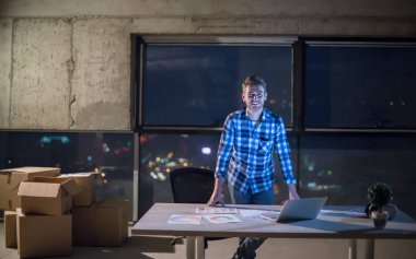 İnşaat alanındaki genç erkek mühendisin portresi gece yeni açılan ofiste dizüstü bilgisayarla belge ve iş akışını kontrol ediyor.