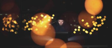 Genç yetenekli hacker karanlık ofiste çalışırken dizüstü bilgisayar kullanıyor. Arka planda büyük şehir ışıkları var.
