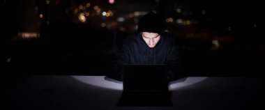 Genç yetenekli hacker karanlık ofiste çalışırken dizüstü bilgisayar kullanıyor. Arka planda büyük şehir ışıkları var.