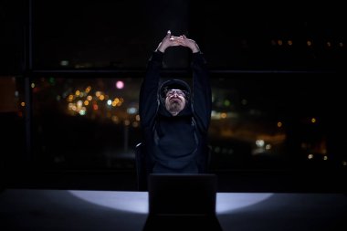 Genç yetenekli hacker karanlık ofiste çalışırken dizüstü bilgisayar kullanıyor. Arka planda büyük şehir ışıkları var.