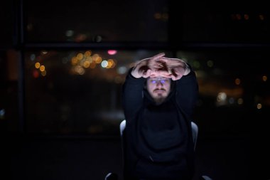 Genç yetenekli hacker karanlık ofiste çalışırken dizüstü bilgisayar kullanıyor. Arka planda büyük şehir ışıkları var.